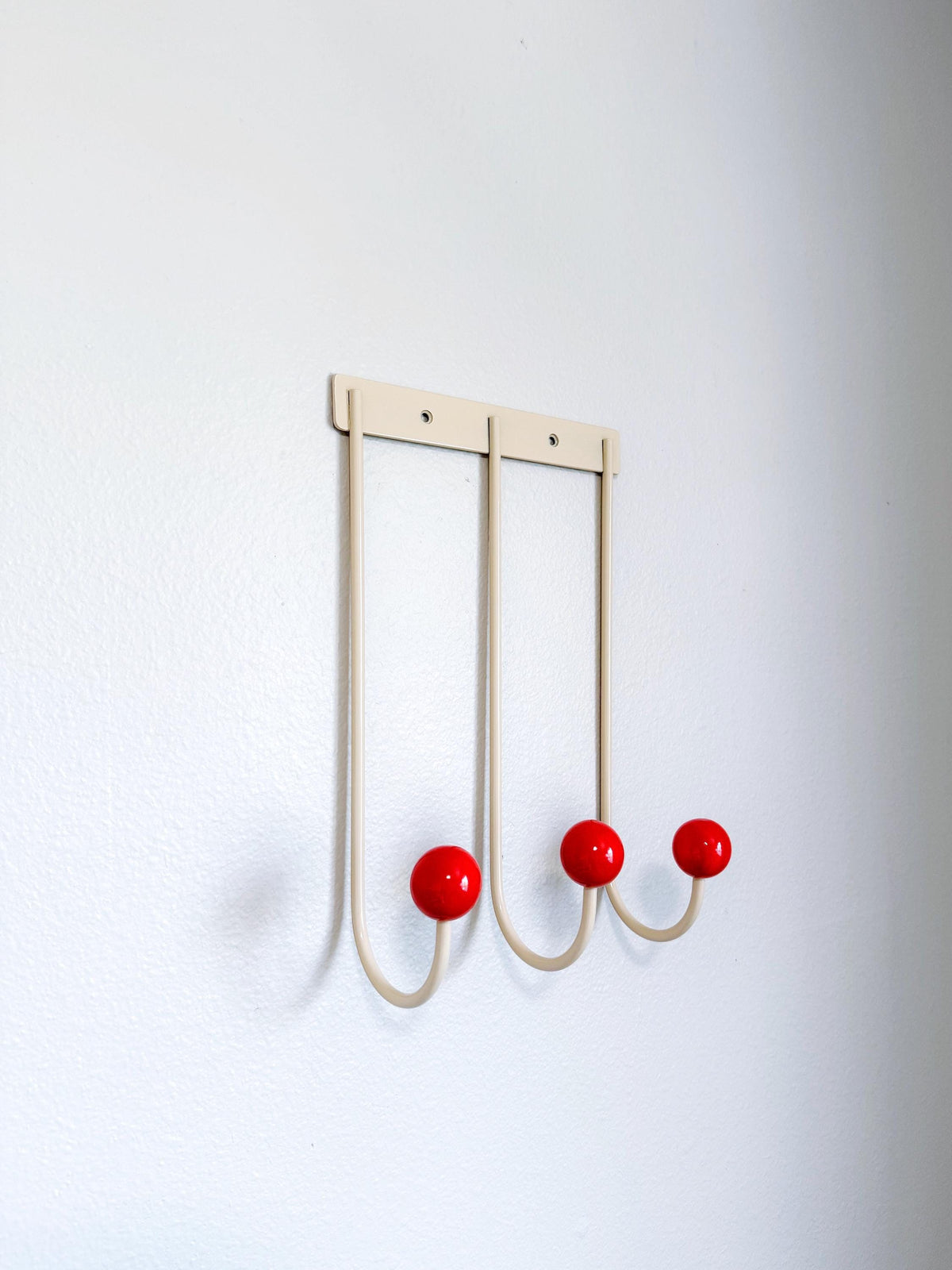 Angled view of a cream wall rack with three long curved hooks and red ball tips.