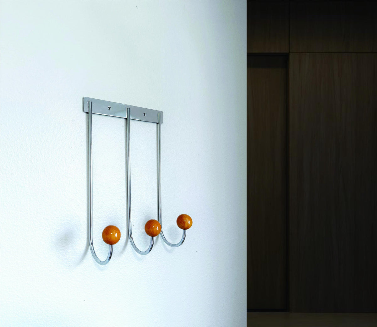 Silver-colored wall rack with three curved hooks and golden-brown ball tips, angled view beside dark paneling.